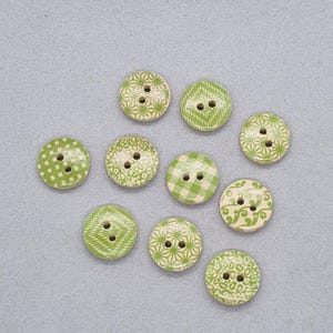 May include: A collection of ten round wooden buttons with two holes each. The buttons feature various patterns in shades of green and white, including geometric shapes, checks, and floral designs. The buttons are arranged on a light gray surface.