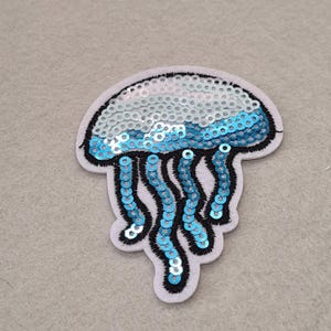 May include: A jellyfish-shaped iron-on patch featuring iridescent white and bright blue sequins. The design is outlined with black embroidery thread on a light gray felt backing, showcasing a rounded bell top and several dangling tentacles.