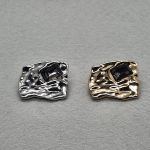 May include: Two square-shaped decorative buttons. One is silver-toned, the other is gold-toned. Each button has a textured surface and a dark gray rectangular gem in the center. The buttons are displayed on a gray surface.