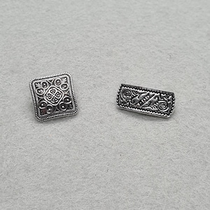 May include: Two silver-coloured metal buttons. One is square with an ornate, symmetrical design. The other is rectangular, also with an intricate pattern. Both buttons have a vintage or antique aesthetic, suitable for sewing or crafting projects.