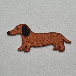 May include: A brown embroidered dachshund patch with a black ear. The dog is shown in profile, with its long body and short legs. The patch is designed for application to clothing or accessories.