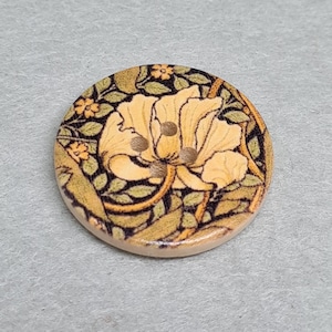 May include: A circular wooden button with a floral pattern. The button showcases a large yellow flower with green leaves and small orange flowers. It has three holes for sewing.
