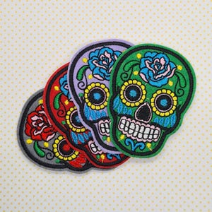 May include: Four embroidered skull patches in various colours. The skulls are in shades of grey, red, purple, and green, each with a floral design. The patches feature yellow, blue, and pink accents, set against a white dotted background.