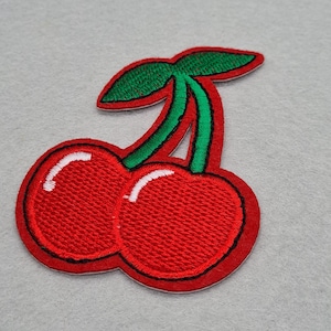 May include: Embroidered patch depicting two vibrant red cherries with green stems and leaves. The cherries have a white highlight and are outlined in black. Suitable for application on clothing or accessories.