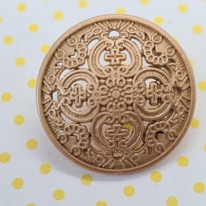 May include: A gold-toned metal button with a decorative, intricate design. The button features a circular shape with a raised, ornate pattern.