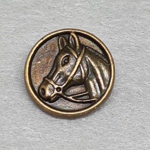 May include: A circular, antique brass button with a detailed horse head profile within a frame. The horse wears a bridle. The button has a vintage, worn appearance, with a diameter of approximately 2.5 cm.