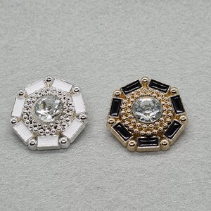 May include: Two decorative buttons with a central clear rhinestone. One button has a silver-tone base with white accents, the other has a gold-tone base with black accents. Both have a geometric design.