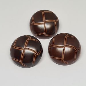 May include: Three dark brown, round buttons with a grid pattern. The buttons have a glossy finish and a raised design, suggesting a vintage style. They are likely for sewing or craft projects. Each button is approximately 2.5 cm in diameter.