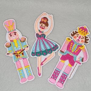 May include: Three embroidered patches featuring two nutcracker figures and one ballerina. The nutcrackers wear pink, blue, and yellow uniforms with crowns. The ballerina wears a teal and pink dress with a yellow hair bow, posed in a dance stance.