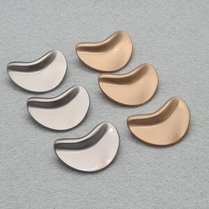 Peut inclure: Six boutons métalliques en forme de rein, dans des tons argent et or. Les boutons ont un design lisse et incurvé et une surface réfléchissante, suggérant une finition polie. Ils sont disposés sur une surface gris clair.