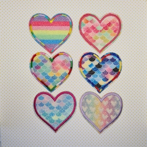 May include: Six heart-shaped patches with different rainbow and mermaid scale patterns. The patches are made of fabric and have a glittery finish.