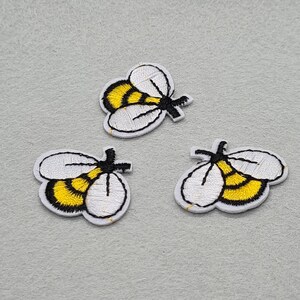 Peut inclure: Trois écussons brodés en forme d'abeille. Chaque écusson présente un corps blanc avec des accents jaunes et noirs. Les abeilles ont un contour noir et sont posées sur un fond gris clair. Idéal pour les vêtements ou accessoires.
