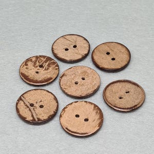 May include: Seven round coconut shell buttons in natural tones. Each button has two sewing holes and displays unique wood grain patterns. The buttons vary in colour, from light tan to medium brown. Ideal for sewing projects.