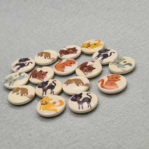 May include: A collection of wooden buttons with cat illustrations. The buttons are off-white with various cat designs in colors like orange, black, brown, and yellow. The cat images are simple and cartoon-like.
