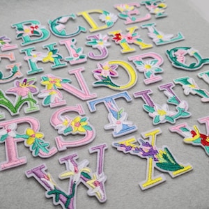 May include: A collection of embroidered alphabet letters, each decorated with floral designs. The letters feature a range of colours, such as pink, yellow, green, and blue. The patches are displayed on a light grey surface, offering a decorative and colourful set.