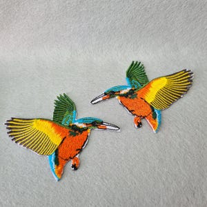 May include: Two embroidered iron-on patches of kingfishers. The birds are facing opposite directions. The patches are orange, yellow, blue, and green.