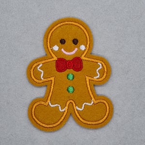 May include: A gingerbread man patch with a smiling face, red bow tie, and green buttons. The patch is outlined in gold and has white accents. Ideal for festive crafts and decorations.