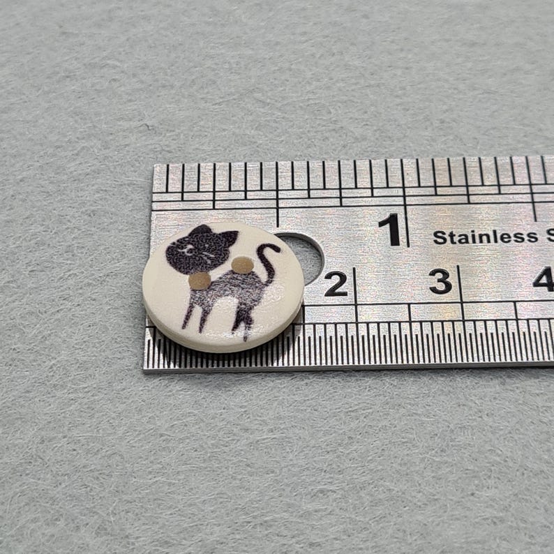 May include: A round, off-white button with a black cat illustration. The cat has a black head, body, and tail, with a tan collar. The button is approximately 0.75 inches in diameter. A stainless steel ruler is next to the button.