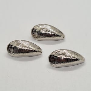 May include: Three shiny, silver-coloured, teardrop-shaped decorative objects. Each has a ridged, shell-like texture. The objects are arranged on a white surface. The design is reminiscent of a mussel shell.