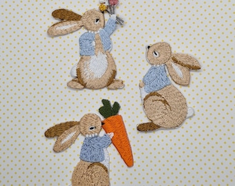 May include: Three embroidered iron-on patches featuring brown rabbits wearing blue shirts. One rabbit holds a bouquet of flowers, another holds a carrot, and the third rabbit is standing with its head tilted up.