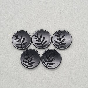 May include: Five black, round buttons with a leaf design. Each button has a two-hole design for sewing. The leaf design is etched into the surface of the buttons.
