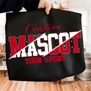 Op de afbeelding: Zwarte draagtas met de tekst "Custom Mascot Your Sport" in rode en witte letters. Het woord "Mascot" wordt doorgesneden door een witte diagonale lijn. De tas wordt door een hand omhoog gehouden.