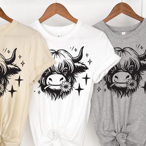 May include: Three t-shirts in beige, white, and gray, each featuring a black and white illustration of a Highland cow with a flower in its mouth. The design includes star accents. The shirts are displayed on wooden hangers.