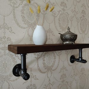 May include: A wooden shelf with black metal pipe brackets. The shelf is mounted to a wall with a patterned wallpaper. A white vase with dried flowers and a silver sugar bowl are on the shelf.