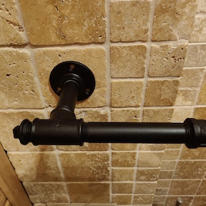 May include: Black metal pipe toilet paper holder mounted on a tiled wall. The holder is in the shape of a T with a round flange on the top.