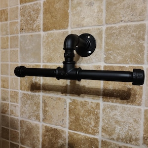May include: Black metal pipe towel rack mounted on a tiled wall. The rack has a simple design with a straight bar and a curved pipe on the left side.