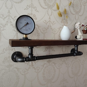 May include: A black metal pipe shelf with a wooden top. The shelf is mounted to a wall and has a black metal pipe rail. A white gauge with black markings is on the shelf. The gauge reads 0.8 MPa. The shelf also has a white ceramic vase and a small white bird figurine.