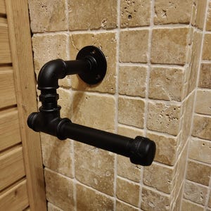 May include: Black metal pipe toilet paper holder mounted on a tiled wall. The holder has a round base and a straight arm.