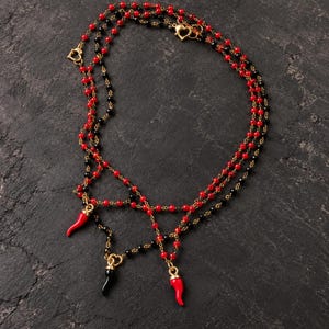 Può includere: Una collana a più fili con perline rosse e nere alternate su una catenina color oro. Il design presenta tre ciondoli pendenti a forma di corno, rossi e neri, arricchiti da piccoli connettori e chiusure color oro a forma di cuore.