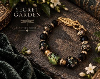 May include: A bracelet featuring black faceted beads, floral-patterned glass beads, a single green textured bead, and gold-toned spacers. The piece is secured with a gold-toned toggle clasp shaped like a feather, accented with small clear crystals.
