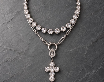 May include: A silver-toned layered necklace featuring a top strand of round clear crystals and a lower chain strand with a central circular clasp. A teardrop-shaped charm and a cross pendant, both encrusted with clear crystals, hang below. VOTIVE is printed.