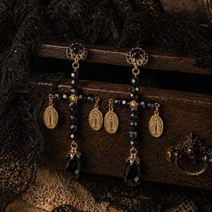 Puede incluir: Pendientes de cruz negros y dorados con cuentas negras y detalles de lágrima. Cada pendiente presenta un medallón dorado con una figura religiosa. Los pendientes se exhiben sobre una superficie de madera con un fondo de encaje oscuro.