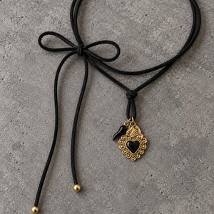 Op de afbeelding: Een zwarte koordketting met een goudkleurige hartvormige hanger met een zwart middenstuk, een kleine zwarte hoornbedel en een decoratieve strik. De uiteinden van het koord zijn afgewerkt met kleine goudkleurige kraaltjes voor een verstelbaar effect.
