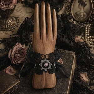 May include: A black lace bracelet featuring a butterfly-shaped design with a central circular medallion containing a small pink floral motif, accented by silver-toned beads and thin black ribbon ties, displayed on a wooden hand model.