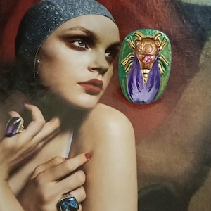 Peut inclure: Une broche en forme de scarabée dorée et violette sur fond vert. Le scarabée a un bijou rose sur sa poitrine.