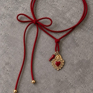Op de afbeelding: Een rode koordketting met een goudkleurige hanger met een centraal rood hart, omringd door filigraan details. Een kleine rode chilipeperbedel hangt naast het hart. Het koord is gestrikt tot een lus en afgewerkt met goudkleurige kraaltjes.