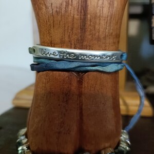 May include: A silver bracelet with a blue and green leather band. The bracelet has the words "live the moment" engraved on it.