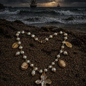 Könnte beinhalten: Eine Perlenkette mit goldenen Akzenten, darunter ein Kreuzanhänger und Kamee-Charms, liegt auf Sand. Die Kette ist herzförmig angeordnet. Der Hintergrund zeigt eine Meereslandschaft mit dunklem, stürmischem Himmel und einem Schiff am Horizont.