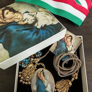 Może przedstawiać: Białe pudełko z pokrywką przedstawiające obraz matki i dziecka. Wewnątrz złoty naszyjnik z wisiorkiem przedstawiającym podobny obraz i pleciony naszyjnik ze sznurka. Na wierzchu wstążka w czerwono-biało-zielone paski.