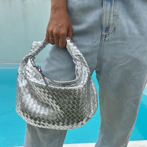 Può includere: Borsa a mano argentata intrecciata con manico annodato e chiusura a zip. La borsa ha una forma arrotondata ed è tenuta su uno sfondo di jeans azzurri e una piscina turchese.