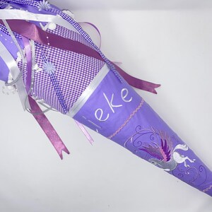 Puede incluir: Un cono de regalo escolar morado con una sección de tela de cuadros blancos, cintas decorativas y pequeños detalles florales. La bolsa está bordada con el nombre "Lieke" en letra blanca y un gráfico de un caballo alado en morado y plata.