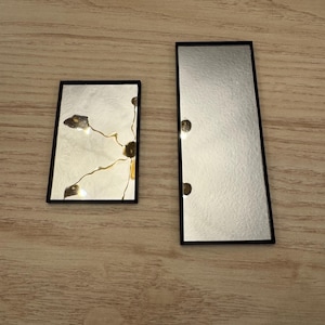 May include: Two rectangular mirrors with black frames rest on a wooden surface. One mirror is smaller and has gold accents, while the other is larger and plain. The mirrors have a reflective surface.