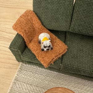 May include: Miniature green sofa with a brown throw pillow. A small figurine of a raccoon with a yellow collar is lying on the pillow. The scene is set on a light wood surface, with a rug and a wooden table.