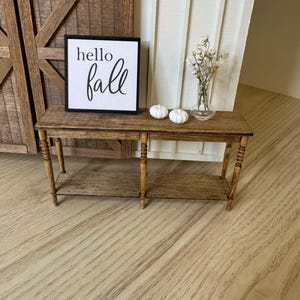 May include: A miniature wooden table with a lower shelf, holding a small sign that reads "hello fall", two white pumpkins, and a glass vase with white flowers. The table is brown with turned legs.