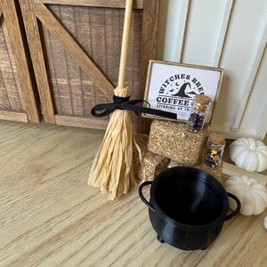 May include: A miniature Halloween scene featuring a small broom with a black ribbon, a black cauldron, and a sign that reads "Witches Brew Coffee". Decorative jars and white pumpkins complete the festive display.