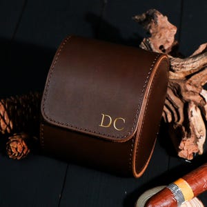 Könnte beinhalten: Ein braunes Leder-Uhrenetui mit goldenen Initialen "DC" auf der Vorderseite. Das Etui ist geschlossen und hat eine gebogene Form. Eine Zigarre liegt auf einer kleinen Holzscheibe neben dem Etui.